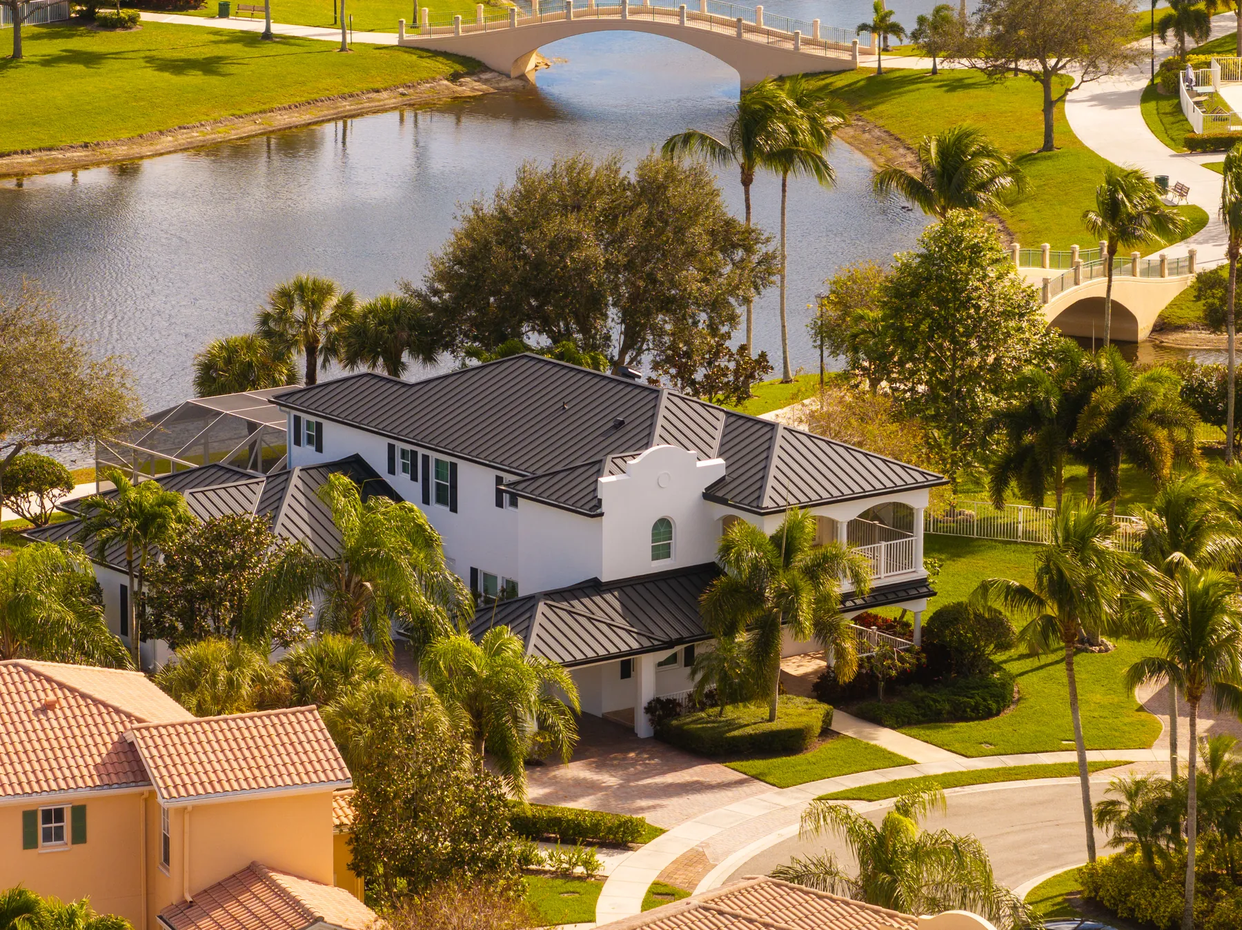 Aerial view of a white house with a dark metal roof beside a lake and a footbridge