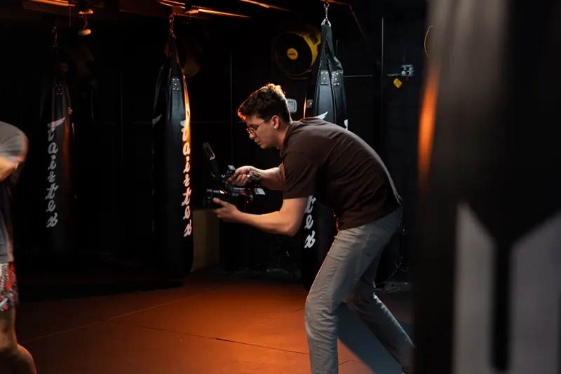A ConnectArt camera operator filming handheld in a fight gym, a heavy bag behind him
