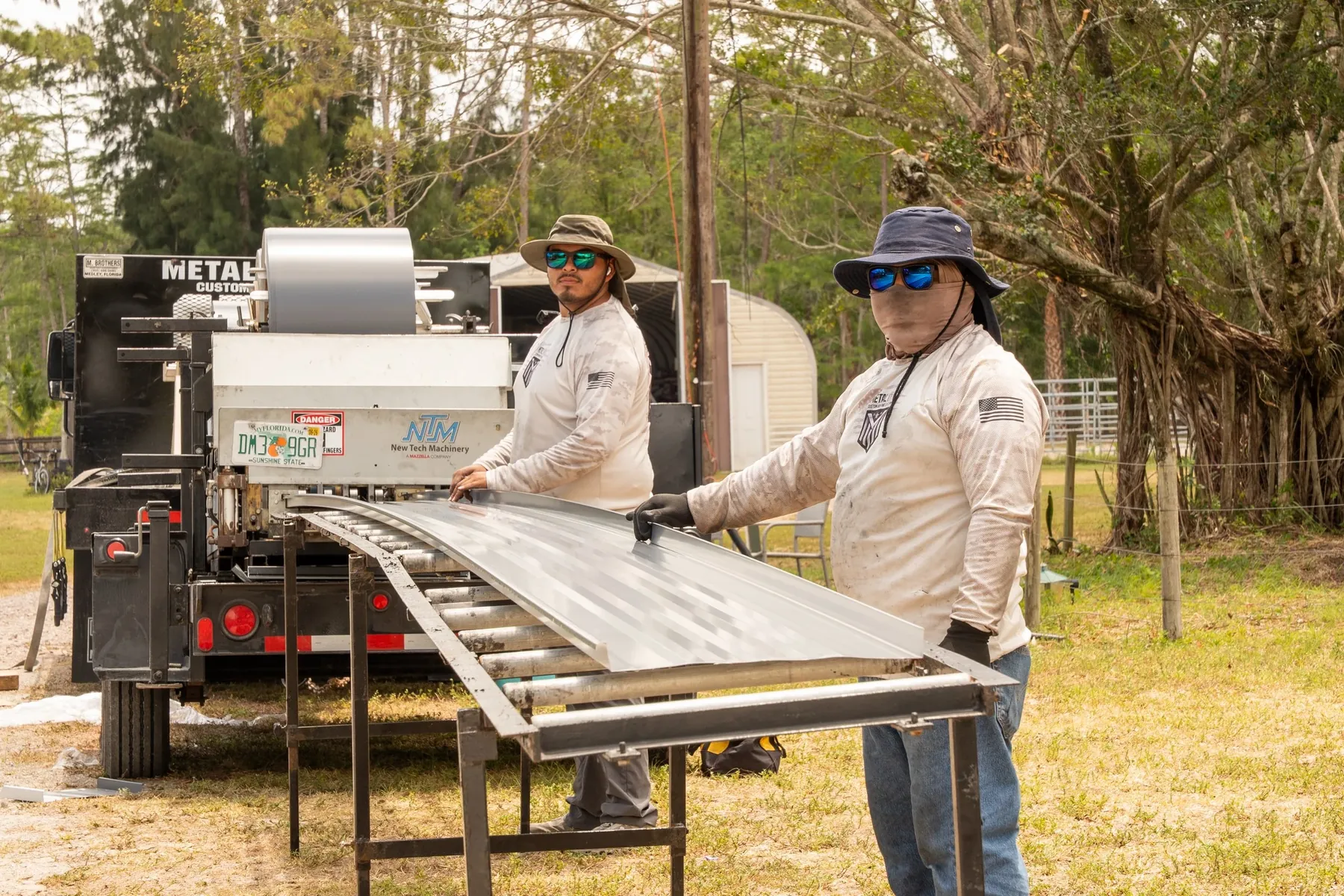 Two workers feeding a metal roofing panel off a forming machine on a trailer