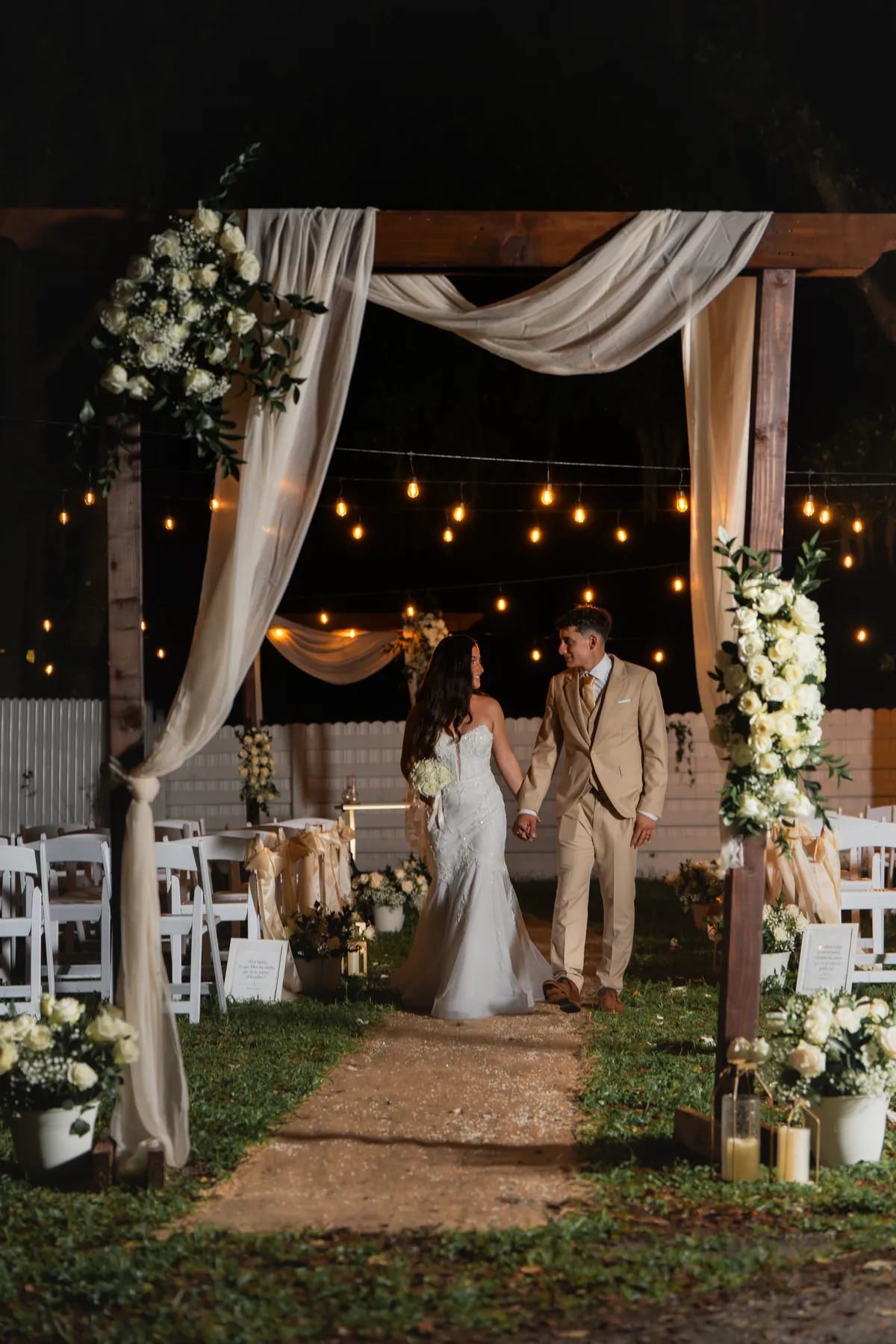 Newlyweds walking up the aisle through a draped wooden arch at night, string lights behind them