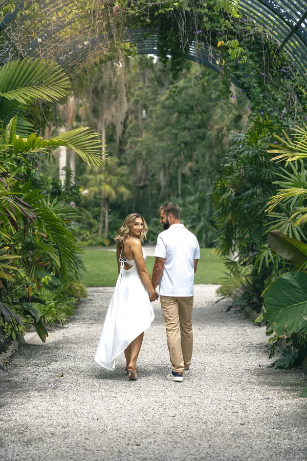 A couple in white walking away from the camera, hand in hand, down a garden path under tropical trees