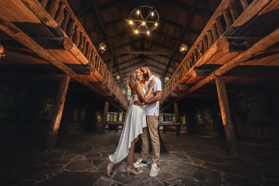 A couple kissing at the far end of a long wooden hall lit warm by hanging lamps