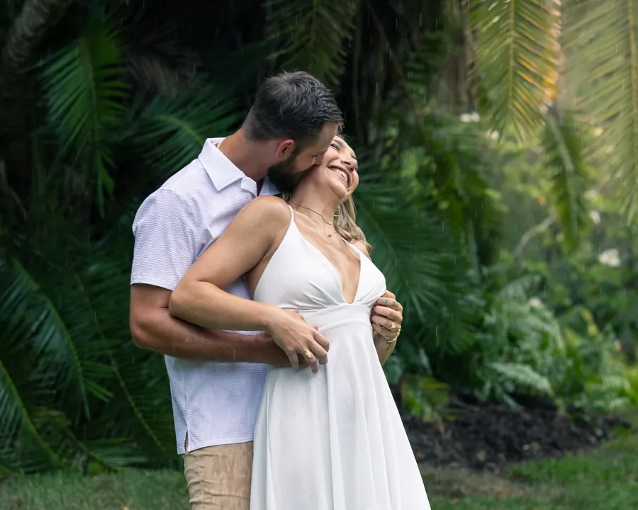 A couple holding each other on the grass under tropical trees, the bride laughing