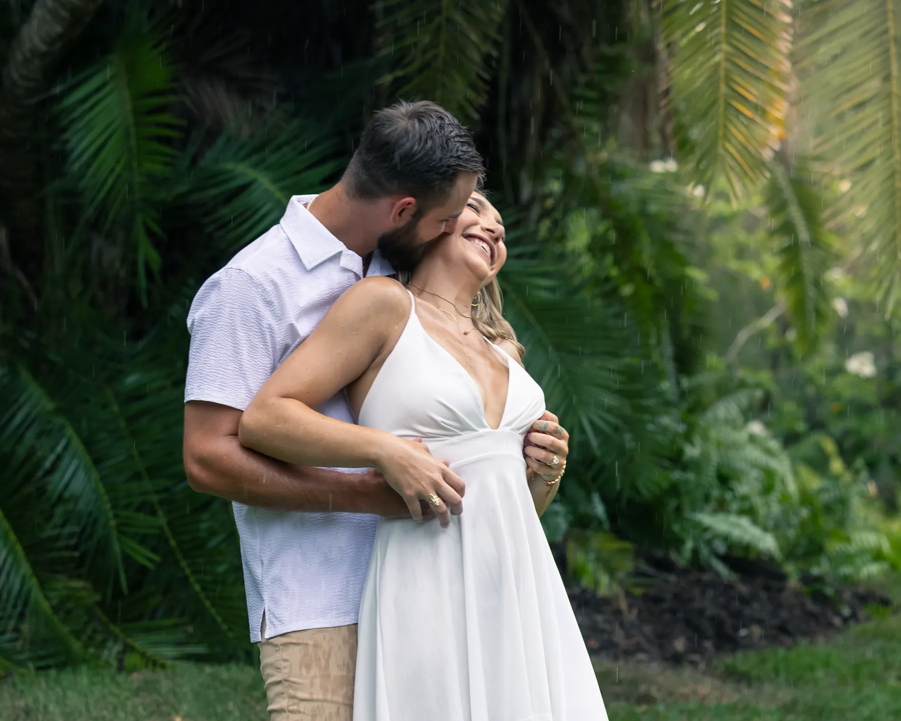 A couple holding each other on the grass under tropical trees, the bride laughing