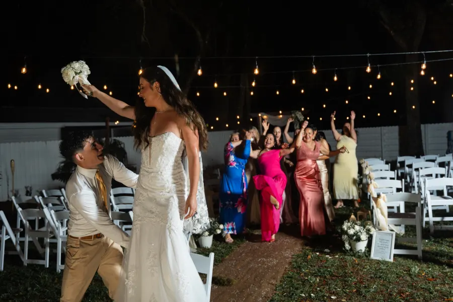 The bride raising her bouquet before the toss while the bridesmaids wait under string lights