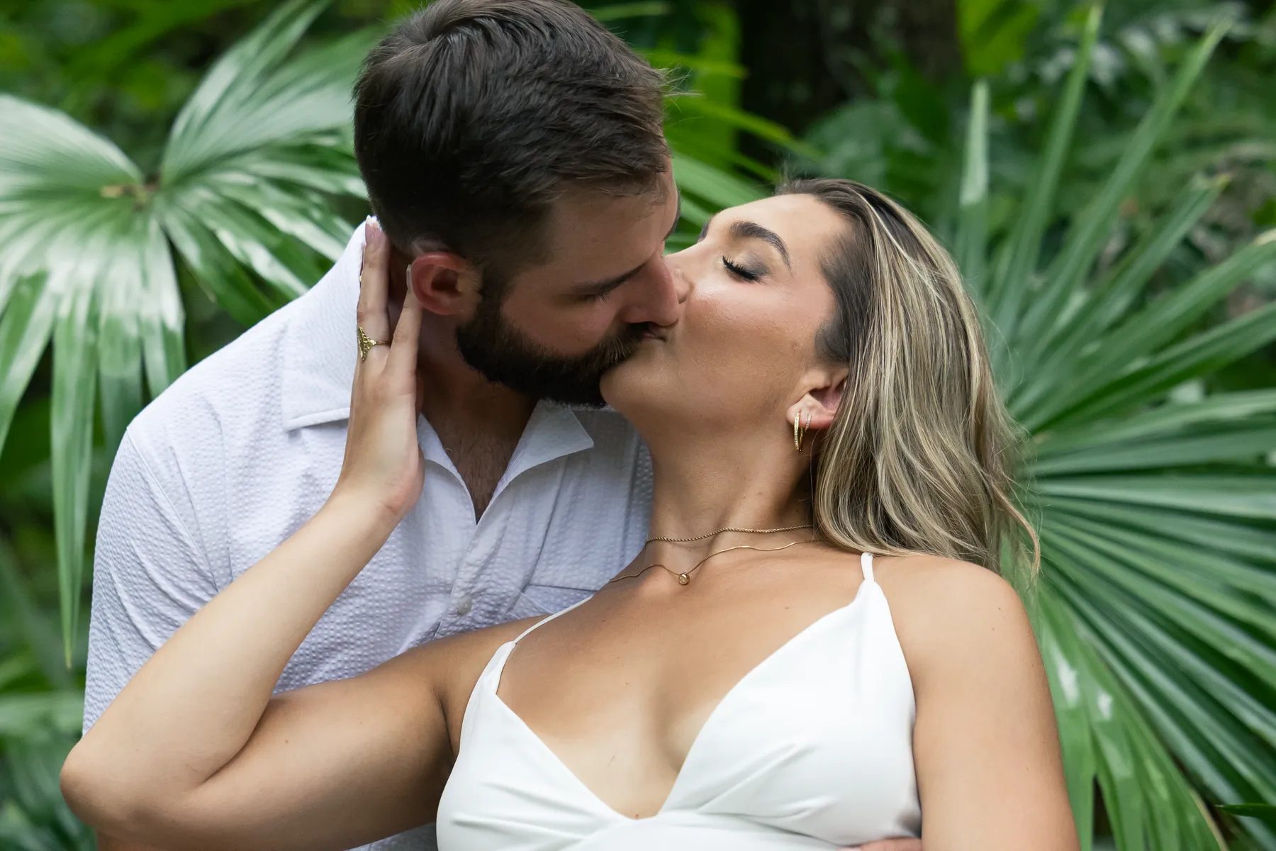 A couple kissing in front of large palm fronds, her hand on his cheek