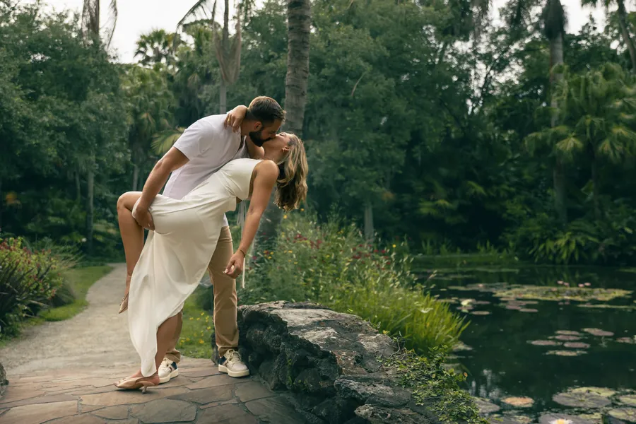 A groom dipping his bride for a kiss on a stone path beside a garden pond