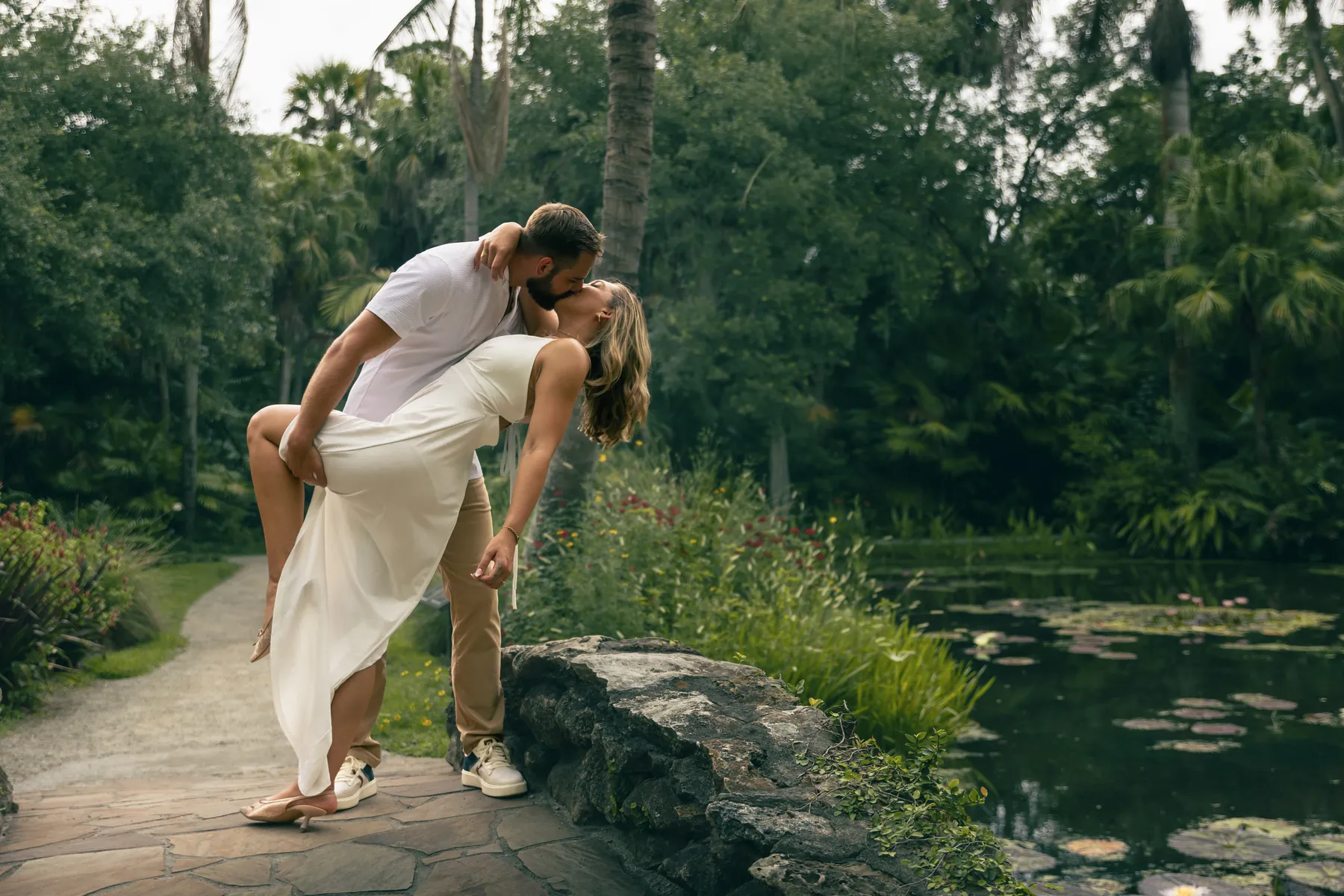 A groom dipping his bride for a kiss on a stone path beside a garden pond