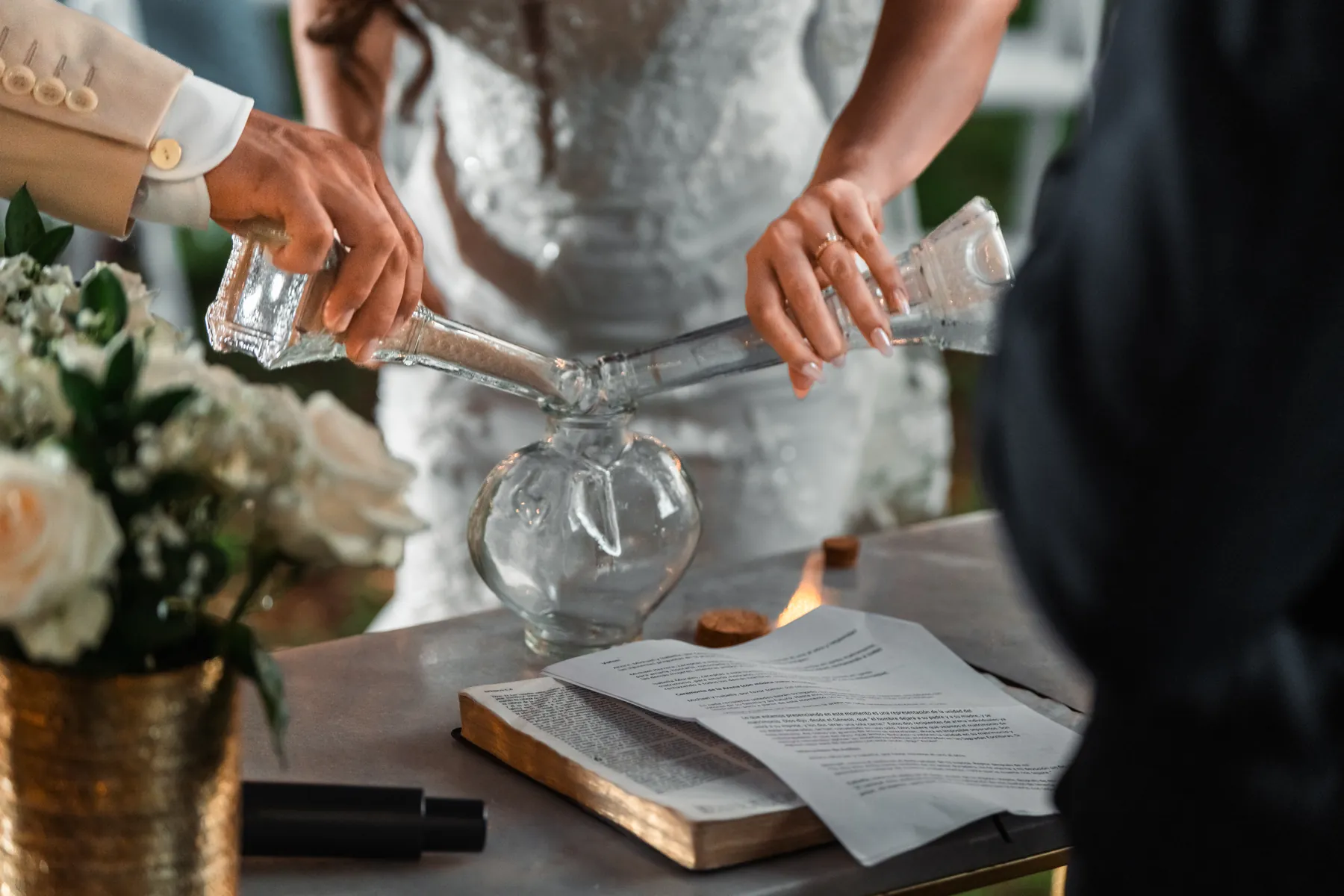 The bride and groom pouring sand into a glass vase during the ceremony, white roses beside them