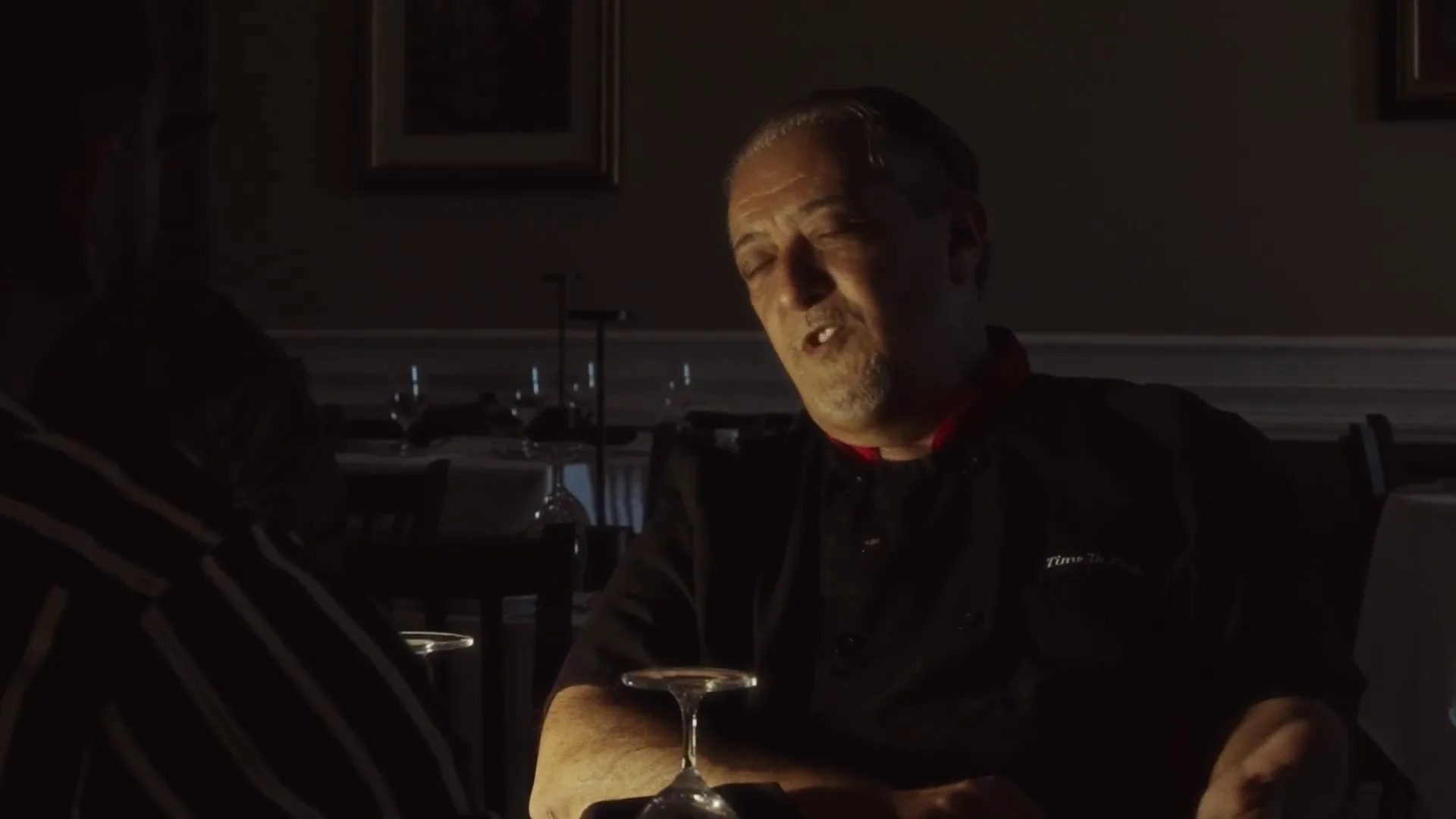 A man at a dinner table in a dark room, a wine glass in front of him