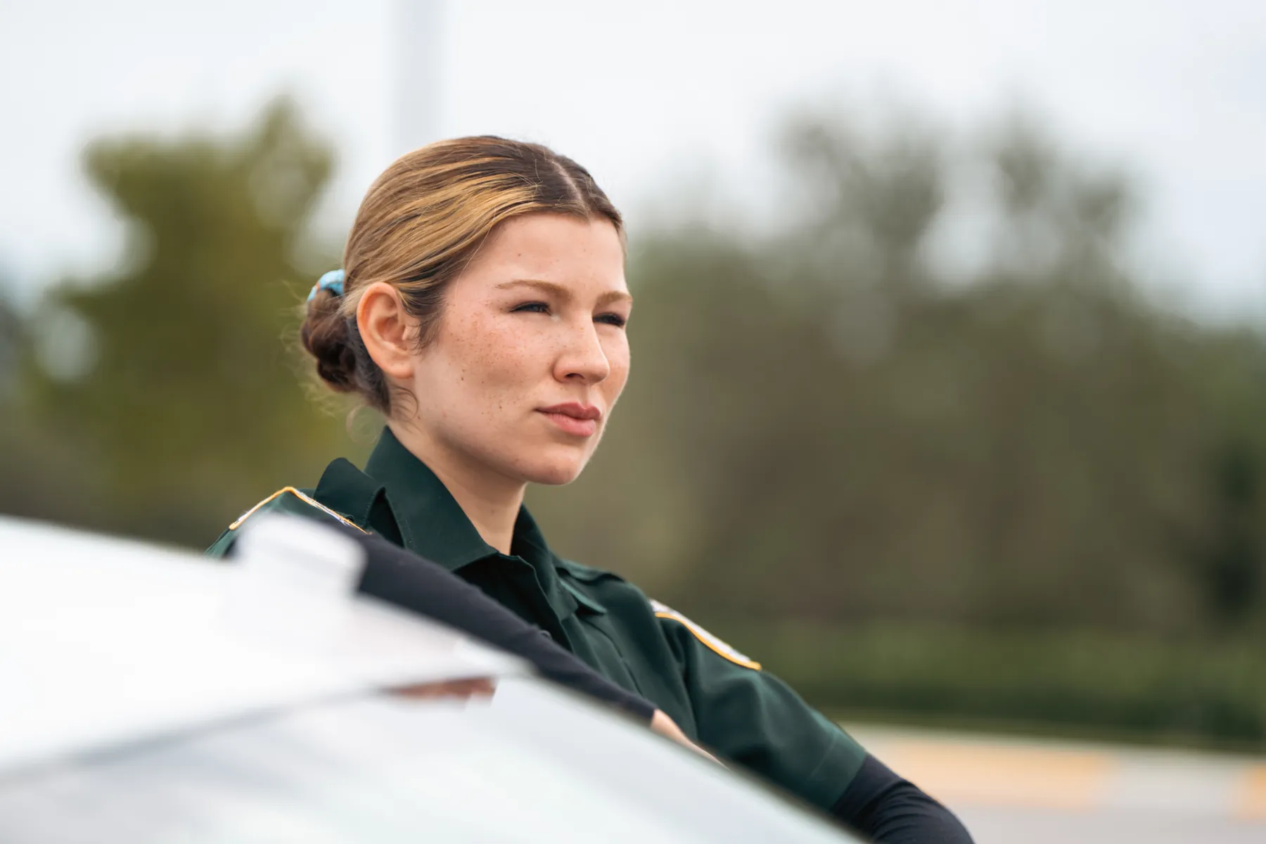 A deputy in uniform leaning on her patrol car, looking off to the side